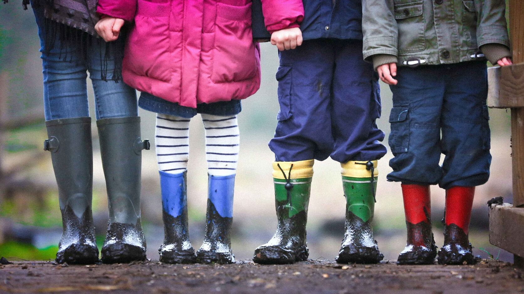 Four children and an adult stand side by side in muddy rubber rain boots — grey, blue, green, and red — photographed from the waist down on a wooden boardwalk.