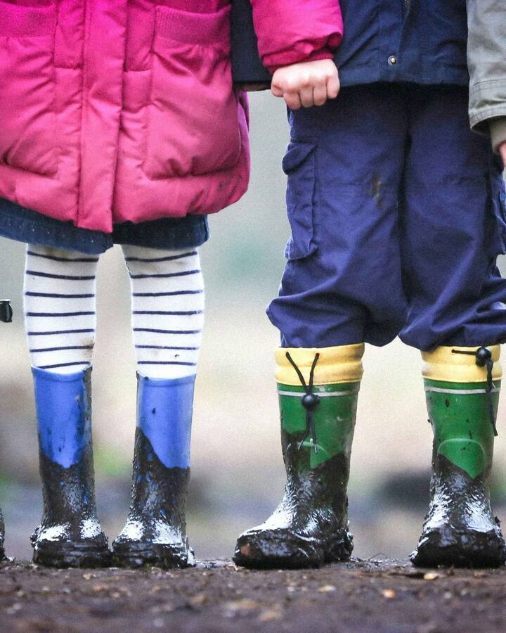 Four children and an adult stand side by side in muddy rubber rain boots — grey, blue, green, and red — photographed from the waist down on a wooden boardwalk.