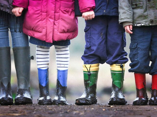 Four children and an adult stand side by side in muddy rubber rain boots — grey, blue, green, and red — photographed from the waist down on a wooden boardwalk.
