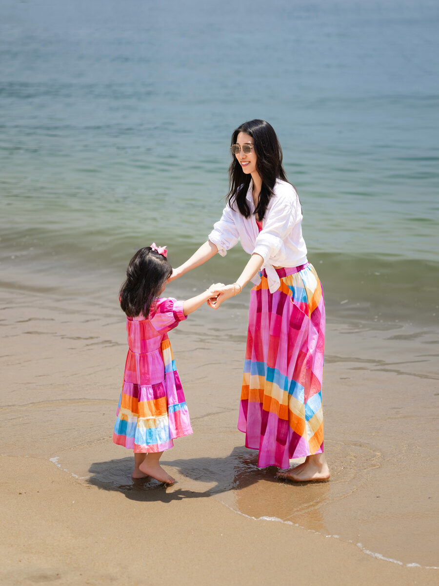 Chris Han and her daughter Luna hold hands in matching rainbow-striped skirts at the water's edge, standing ankle-deep in the shallows.