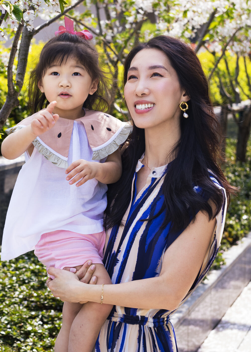 Chris Han holds her daughter Luna under a cherry blossom tree, both looking up as Luna points at the branches.