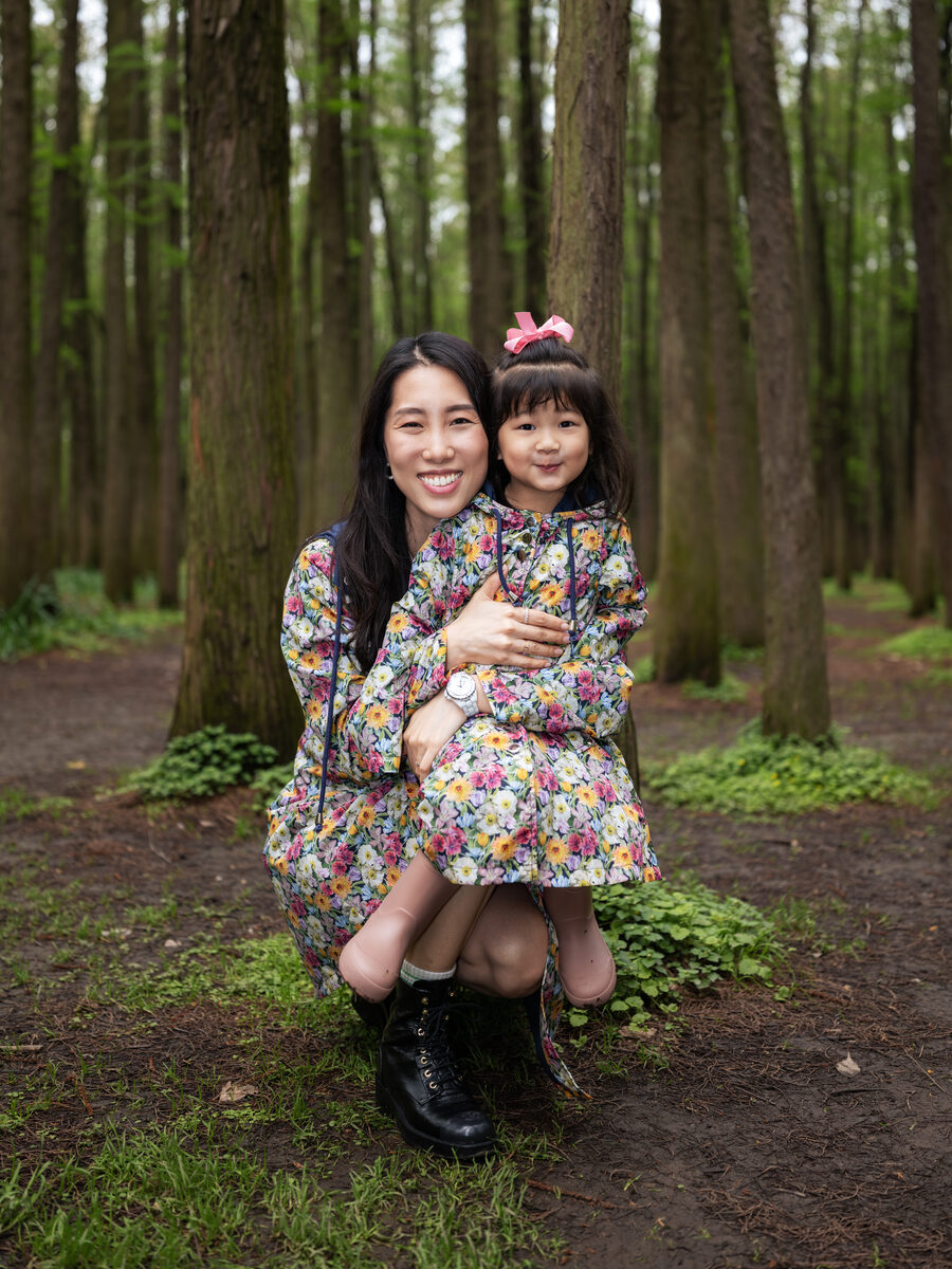 Chris Han crouches on a woodland path holding Luna in front of her, the two of them in matching floral raincoats, smiling at the camera.