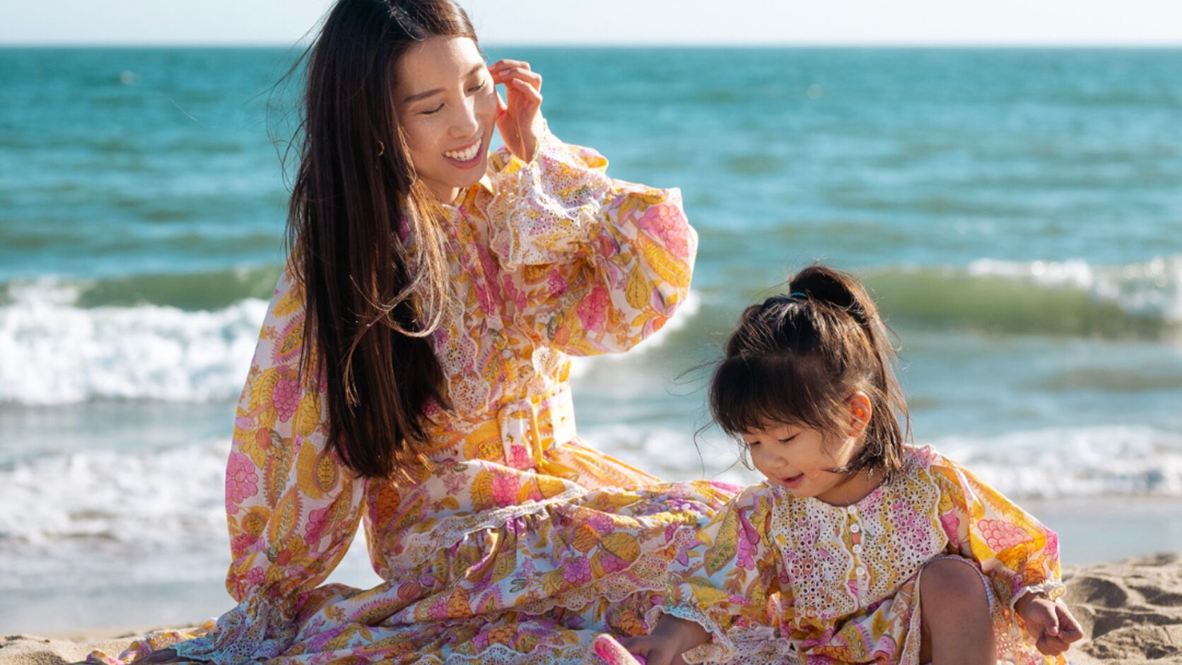 Chris Han sits in the sand in a floral maxi dress, laughing as she tucks her hair behind her ear, while her daughter Luna plays beside her in a matching print with the ocean behind them.