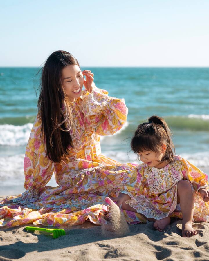 Chris Han sits in the sand in a floral maxi dress beside her daughter Luna, the ocean behind them.