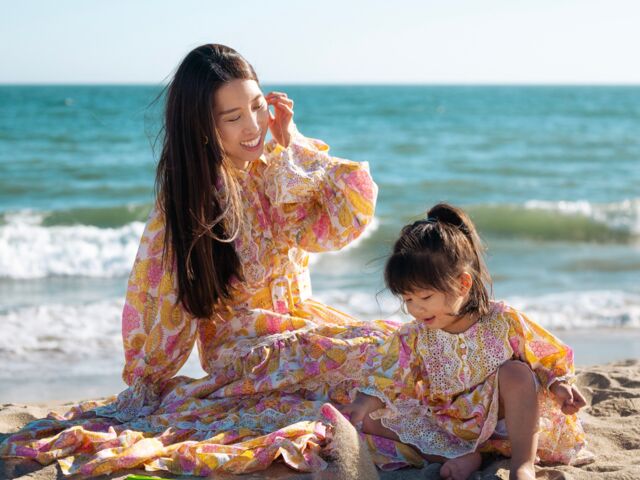 Chris Han sits in the sand in a floral maxi dress beside her daughter Luna, the ocean behind them.