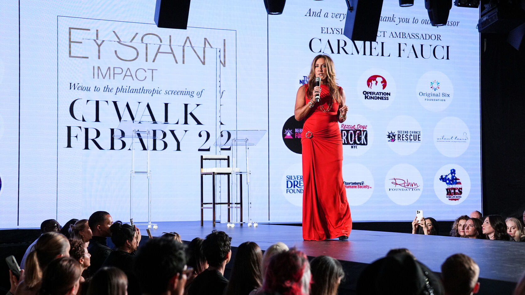 Dr. Christina Rahm, in a floor-length red dress, speaks into a microphone on stage at an Elysian Impact philanthropic screening, sponsor logos including the Rahm Foundation on the screen behind her and the audience in the foreground.