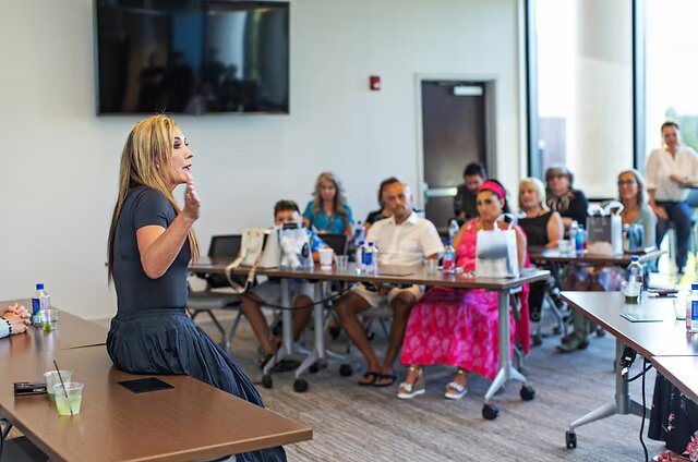 Dr. Christina Rahm, in a navy top and full skirt, perches on the edge of a table as she speaks to a seated group of adults and children in a bright conference room.