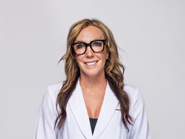 Dr. Christina Rahm, in black square-framed glasses and a white lab coat over a black top, smiles at the camera with her hands in her pockets against a pale grey studio backdrop.