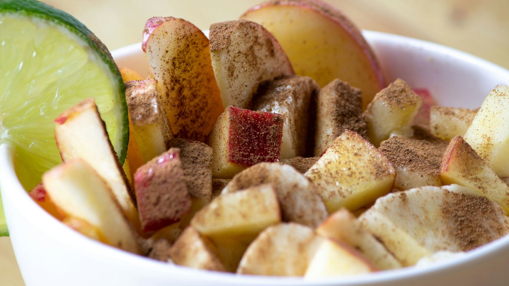 A white bowl of chopped and sliced apple dusted with cinnamon sugar, a wedge of lime tucked against one side, on a pale wooden table.