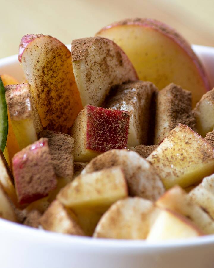A white bowl of chopped and sliced apple dusted with cinnamon sugar, a wedge of lime tucked against one side, on a pale wooden table.