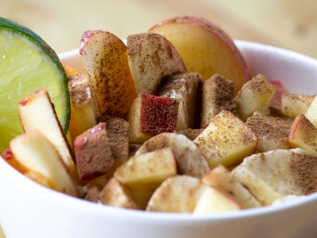 A white bowl of chopped and sliced apple dusted with cinnamon sugar, a wedge of lime tucked against one side, on a pale wooden table.