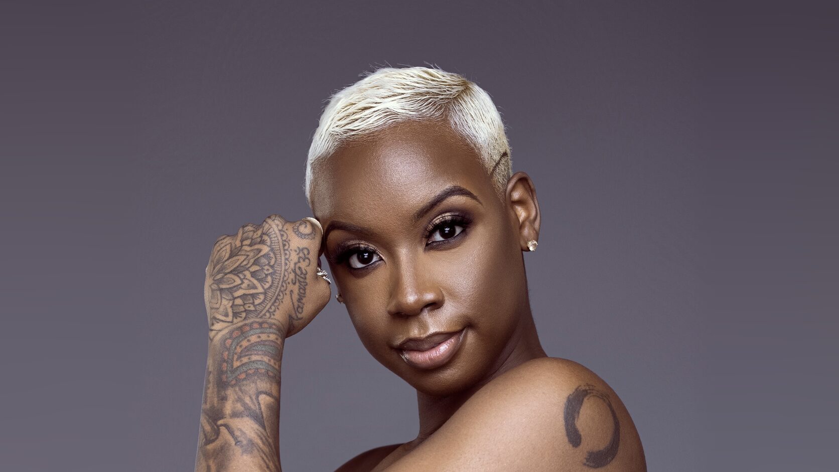 Coach Zha, with a short platinum-blonde cut and a tattooed hand raised to her temple, looks over her bare shoulder at the camera against a soft gray backdrop.
