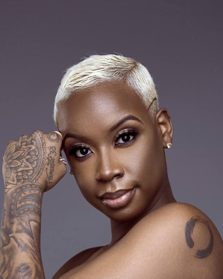 Coach Zha, with a short platinum-blonde cut and a tattooed hand raised to her temple, looks over her bare shoulder at the camera against a soft gray backdrop.