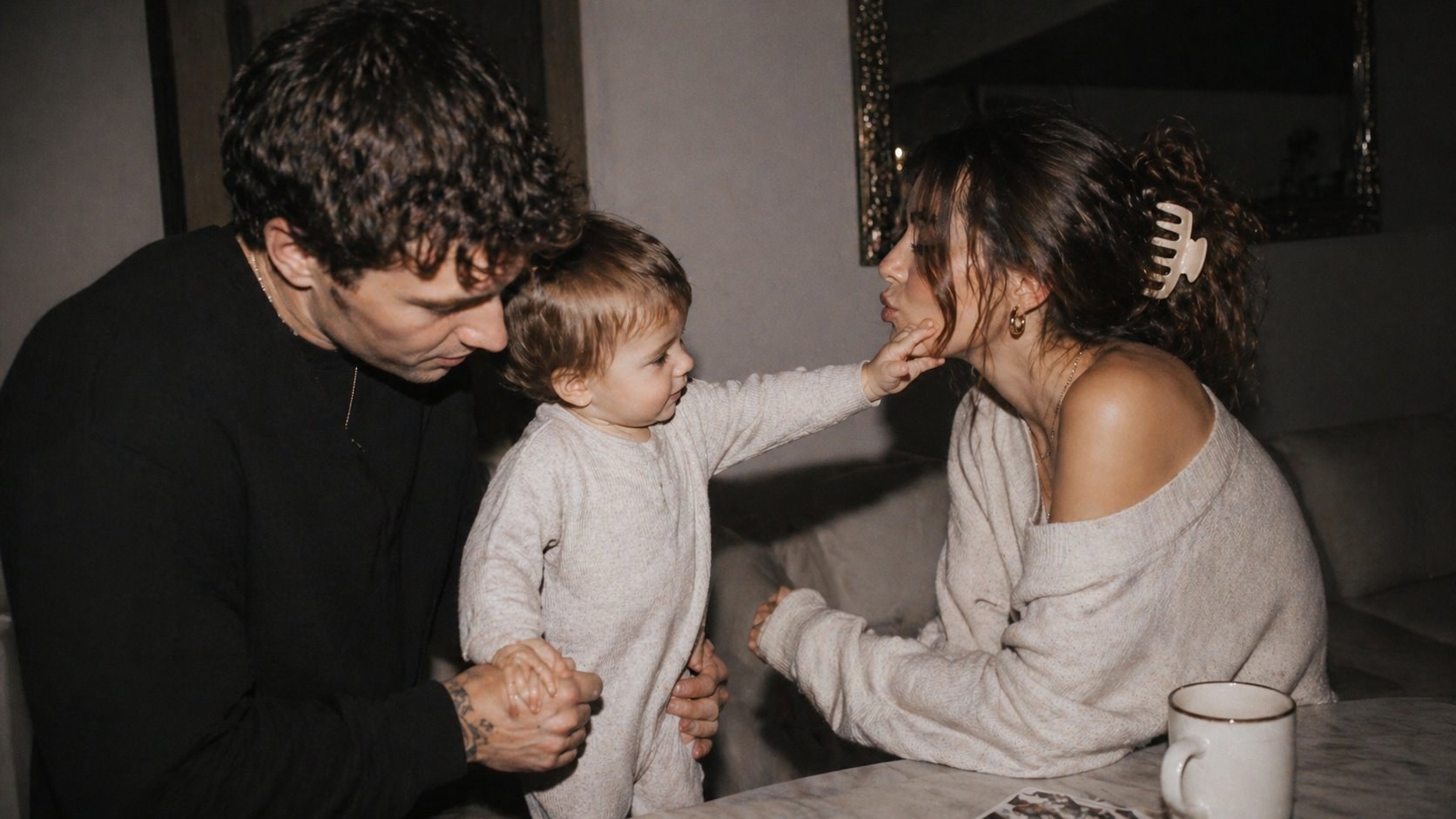 A mother and father with their toddler in a softly lit interior; the child reaches across to touch their mother's face.