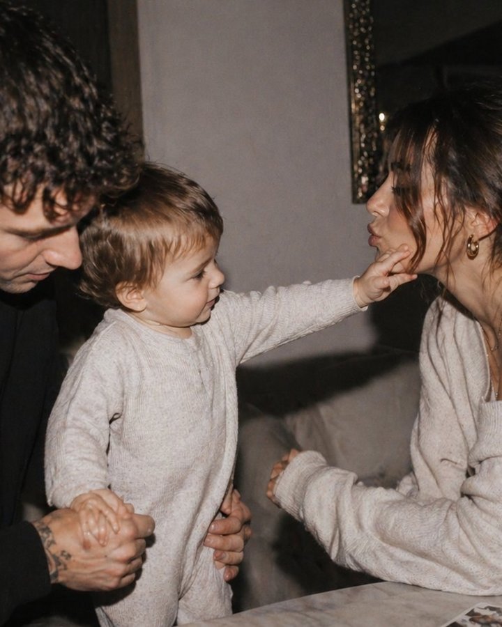 A mother and father with their toddler in a softly lit interior; the child reaches across to touch their mother's face.