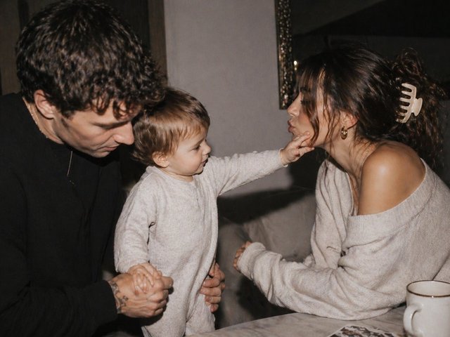 A mother and father with their toddler in a softly lit interior; the child reaches across to touch their mother’s face.