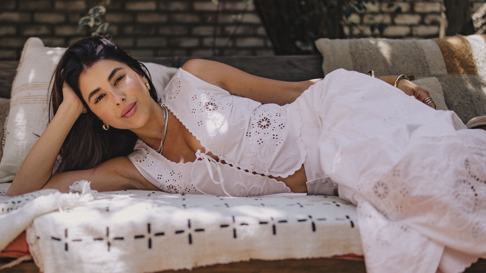 Daniella Monet lies on her side across a cream daybed in a white eyelet top and skirt, her head propped on one hand, gold hoops and layered necklaces catching dappled sunlight against a brick wall.