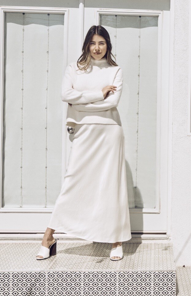 Daniella Monet stands with her arms crossed on a patterned tile step in front of white French doors, wearing a cream turtleneck sweater, a long satin skirt and white mules.