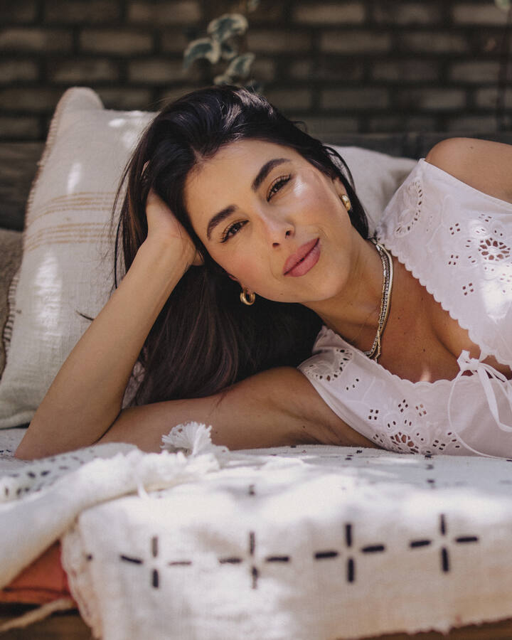 Daniella Monet lies on her side across a cream daybed in a white eyelet top and skirt, her head propped on one hand, gold hoops and layered necklaces catching dappled sunlight against a brick wall.