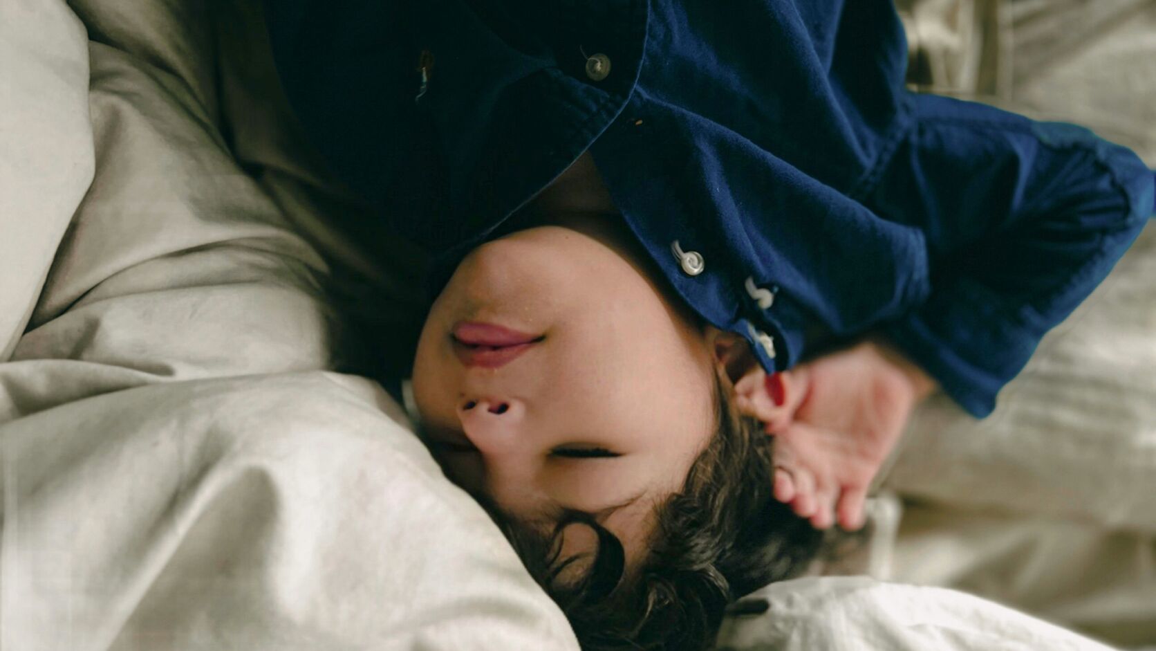 A young child with dark curly hair lies upside down on rumpled cream bedding in a navy button-up shirt, eyes closed and tongue poking out, one hand tucked behind their head.
