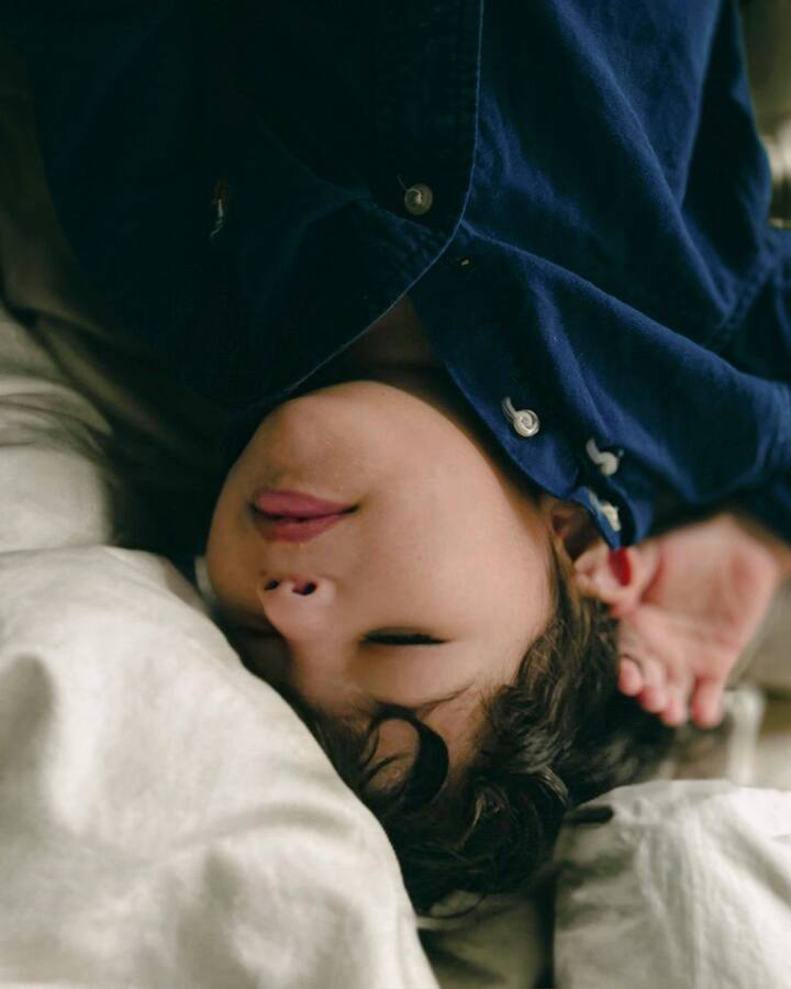 A young child with dark curly hair lies upside down on rumpled cream bedding in a navy button-up shirt, eyes closed and tongue poking out, one hand tucked behind their head.