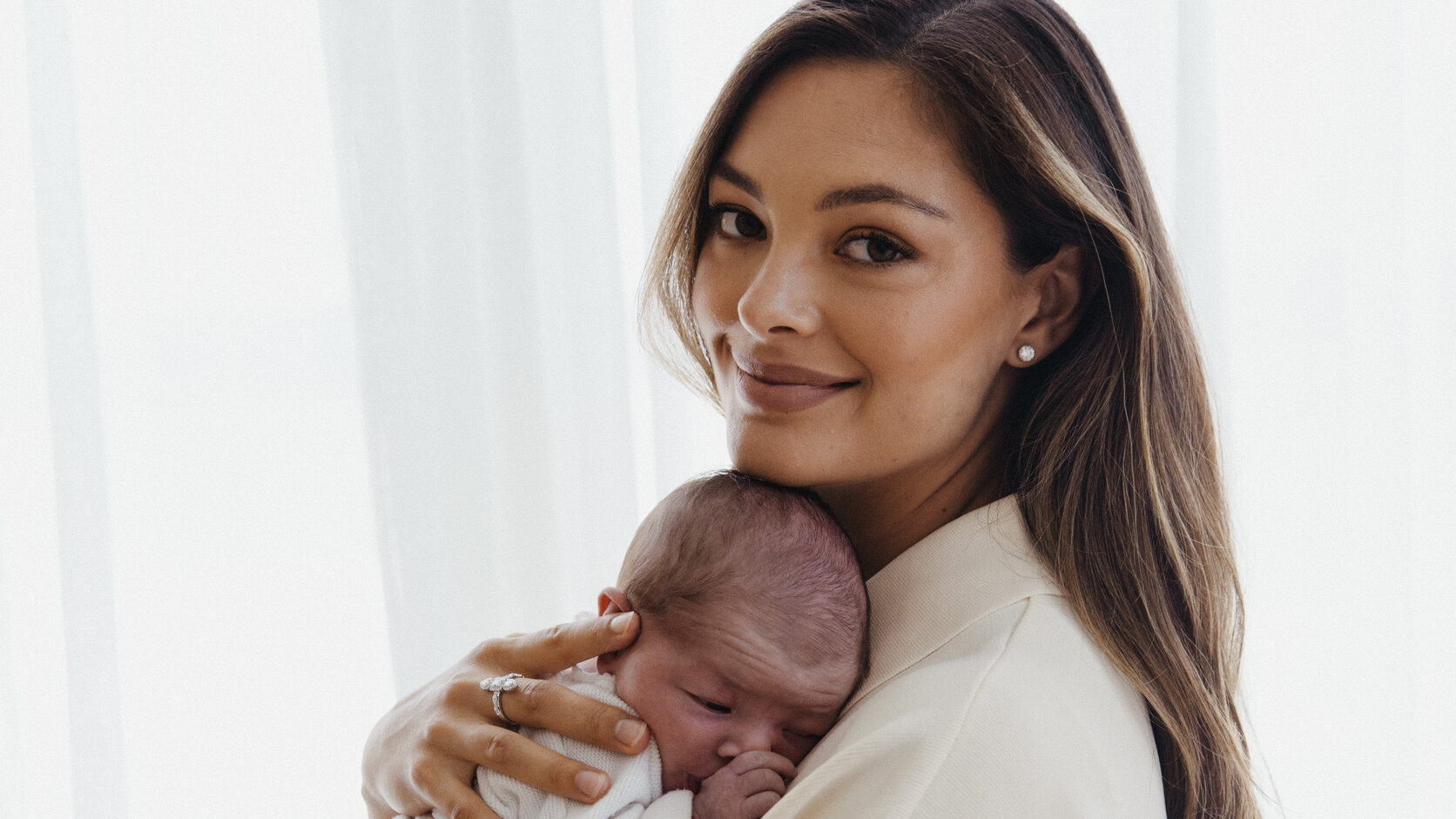 Demi-Leigh Tebow, in a cream jacket, holds her sleeping newborn daughter cheek-to-cheek against a backdrop of sheer white curtains.