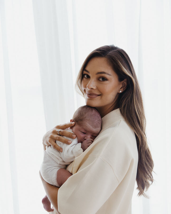 Demi-Leigh Tebow, in a cream jacket, holds her sleeping newborn daughter cheek-to-cheek against a backdrop of sheer white curtains.