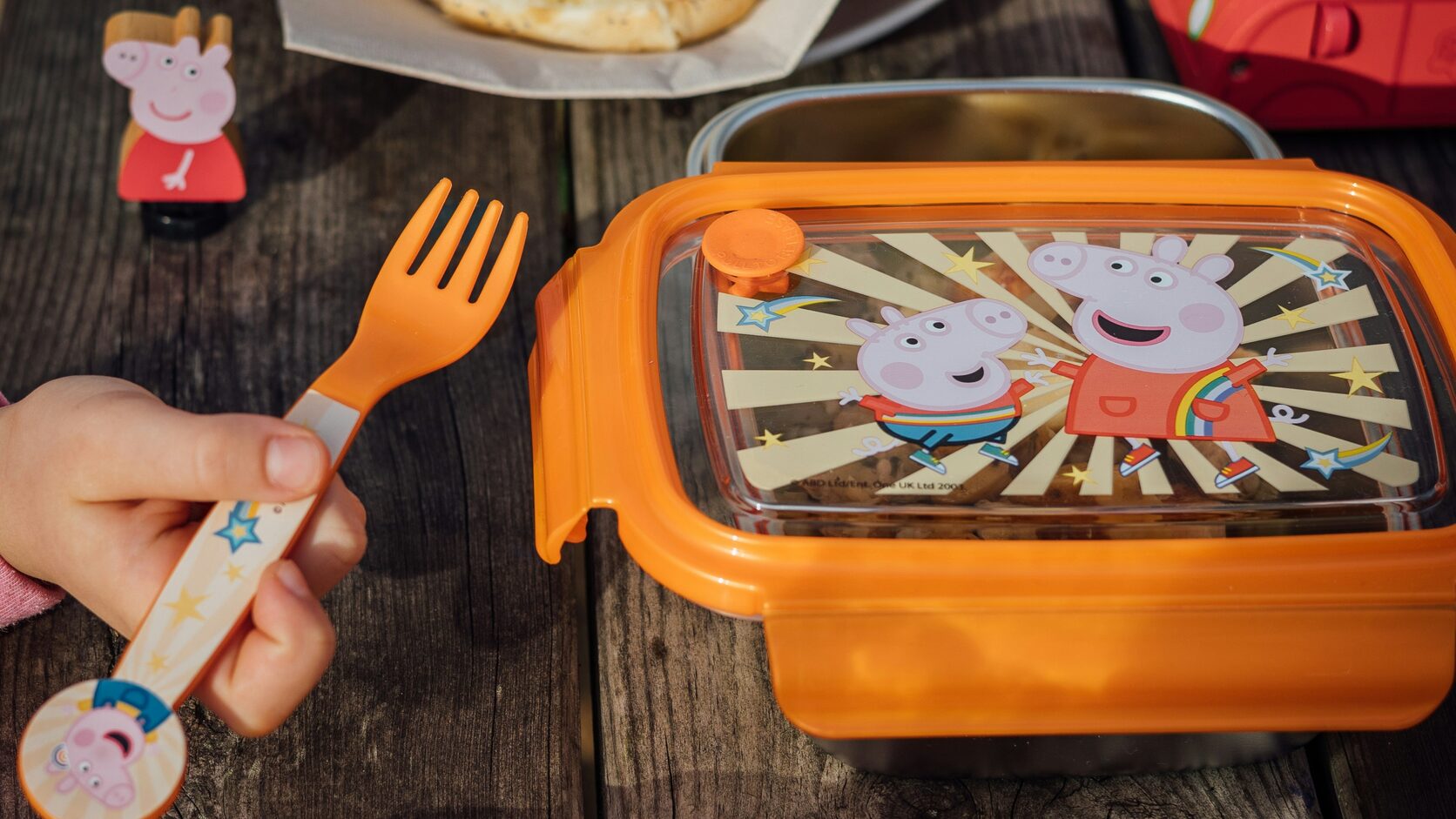 A child's hand reaching for an orange kids' lunch container on a weathered wooden picnic table, a bagel sandwich on a plate beside it.
