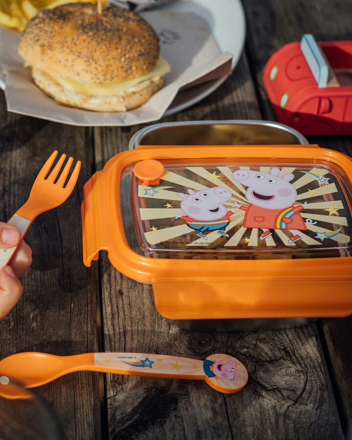 A child's hand reaching for an orange kids' lunch container on a weathered wooden picnic table, a bagel sandwich on a plate beside it.