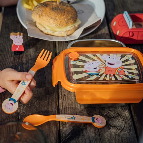 A child's hand reaching for an orange kids' lunch container on a weathered wooden picnic table, a bagel sandwich on a plate beside it.