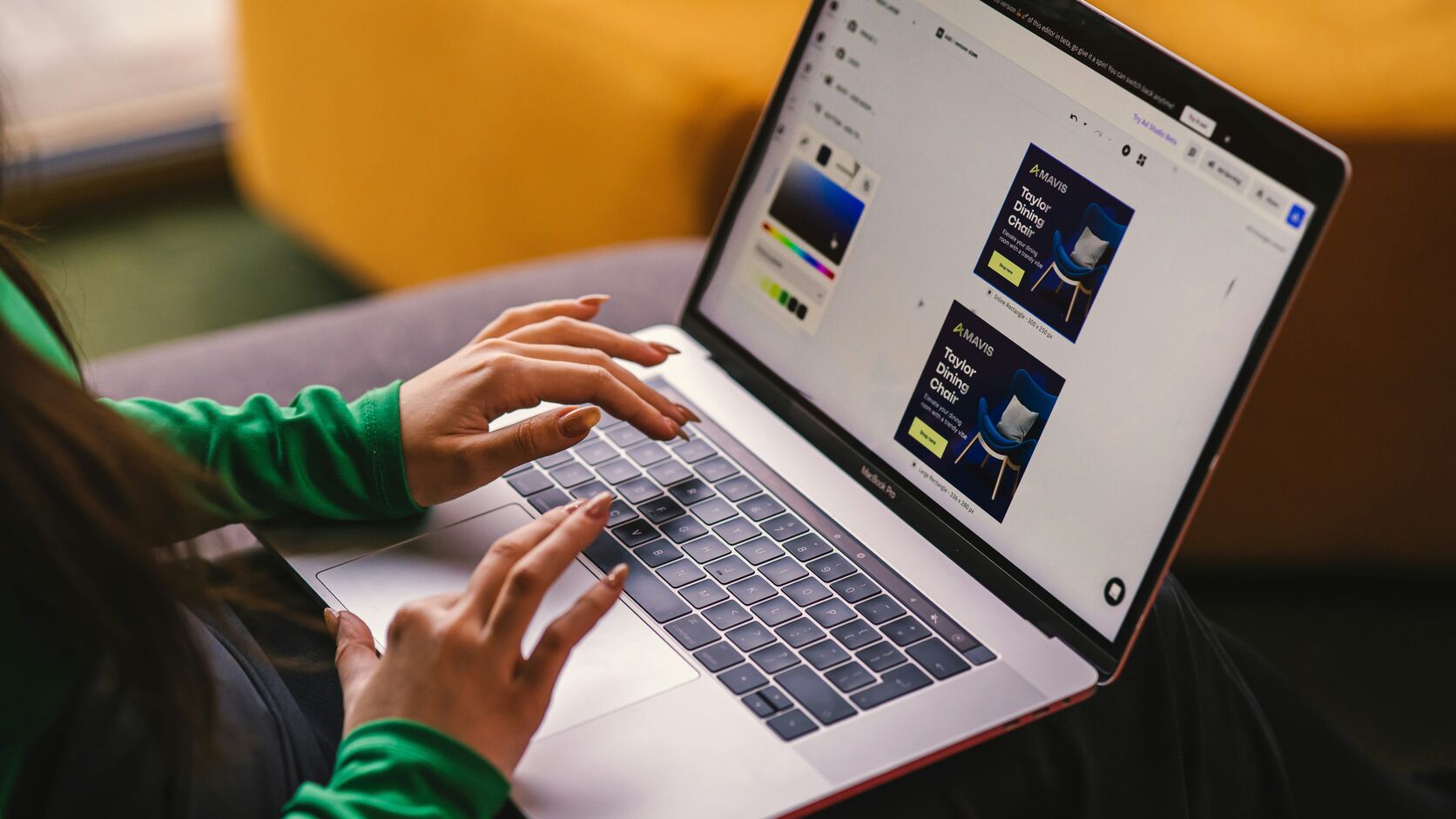 A woman in a green sweater works on a MacBook on her lap, the screen showing an online store's product ad being designed.