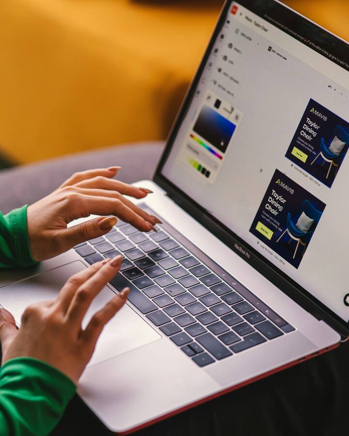 A woman in a green sweater works on a MacBook on her lap, the screen showing an online store's product ad being designed.