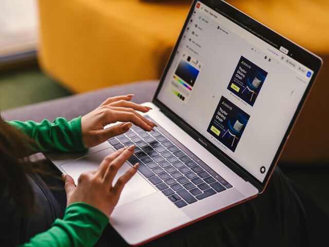 A woman in a green sweater works on a MacBook on her lap, the screen showing an online store's product ad being designed.