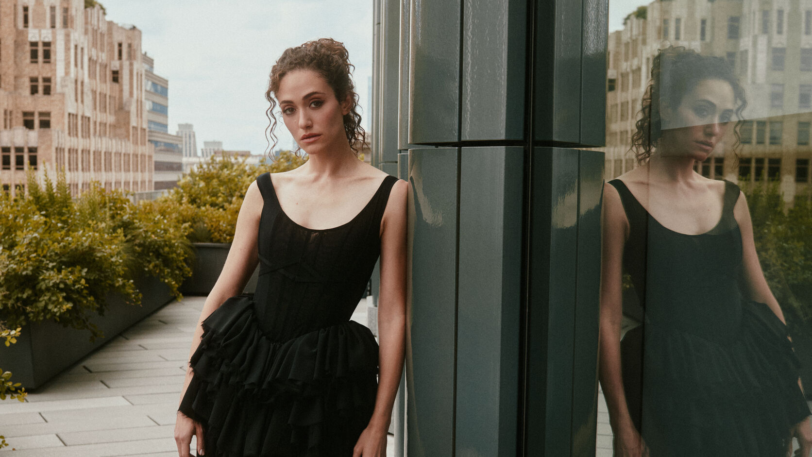 Emmy Rossum leans against a dark glass column on a city rooftop in a black corseted, tiered tulle dress, her reflection beside her.