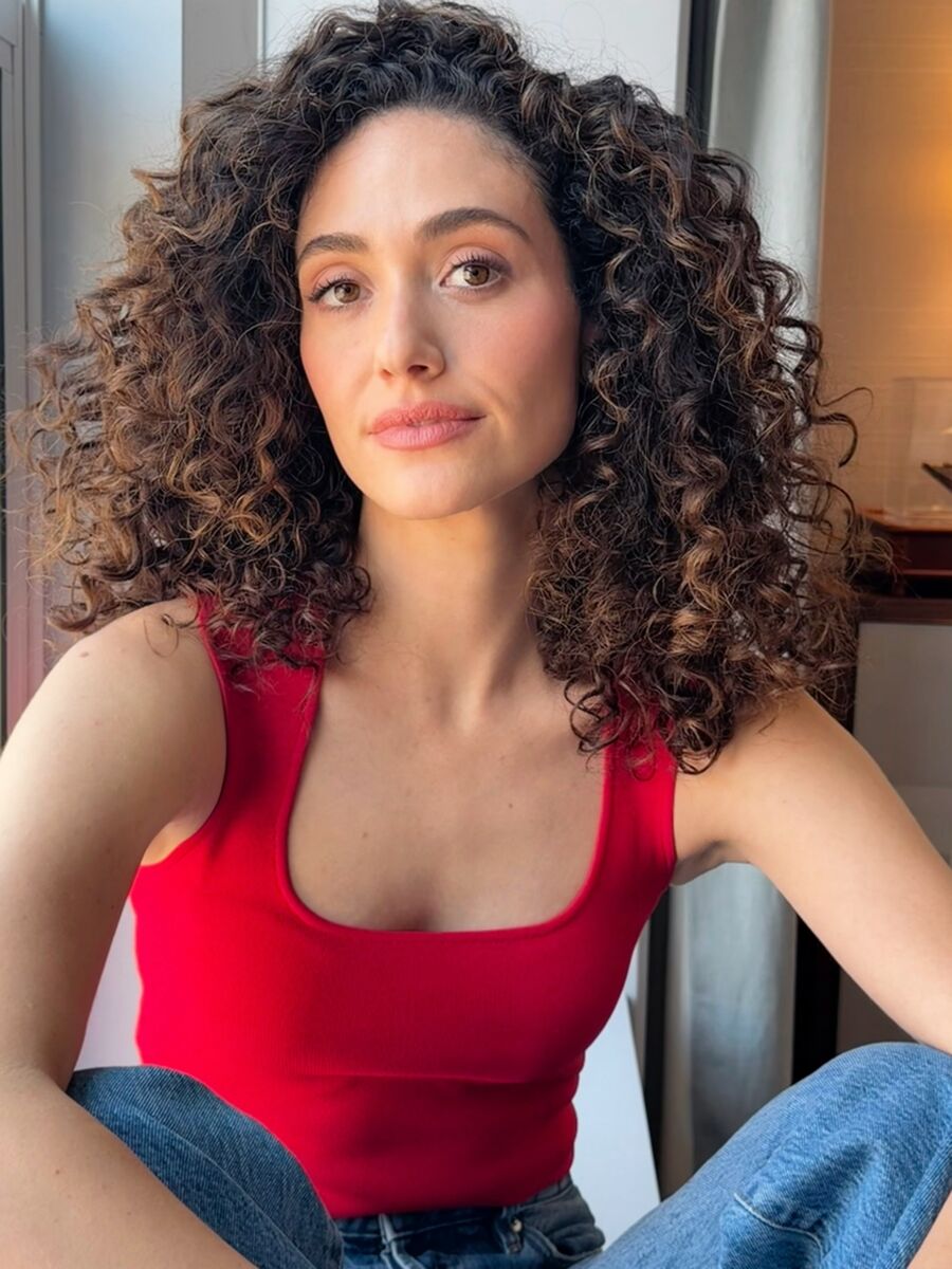Emmy Rossum sits by a window in a red tank top and jeans, her curls loose around her shoulders, in warm afternoon light.