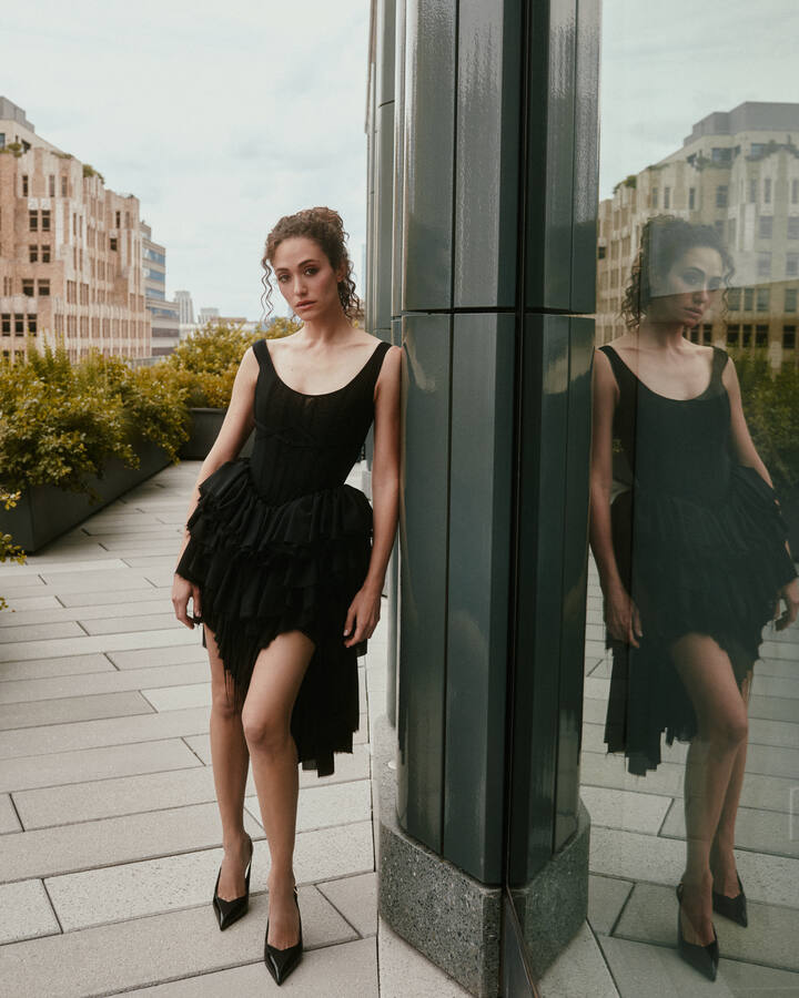 Emmy Rossum leans against a dark glass column on a city rooftop in a black corseted, tiered tulle dress, her reflection beside her.