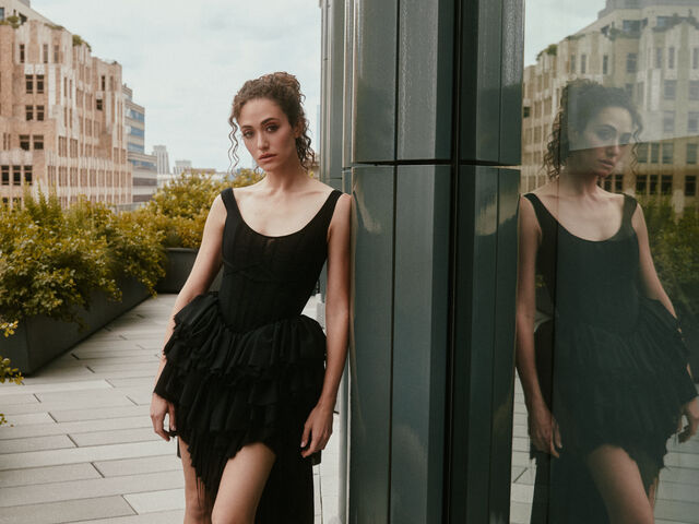 Emmy Rossum leans against a dark glass column on a city rooftop in a black corseted, tiered tulle dress, her reflection beside her.
