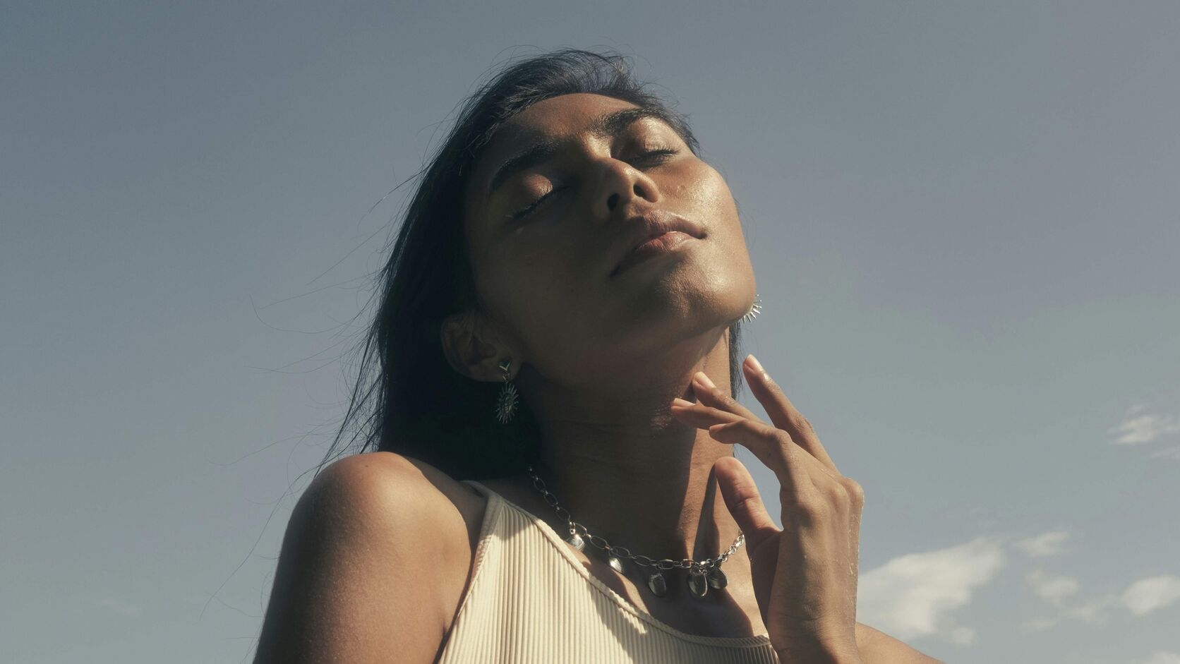 A woman with eyes closed tilts her face up to the sun, one hand resting at her collarbone, against a clear sky.