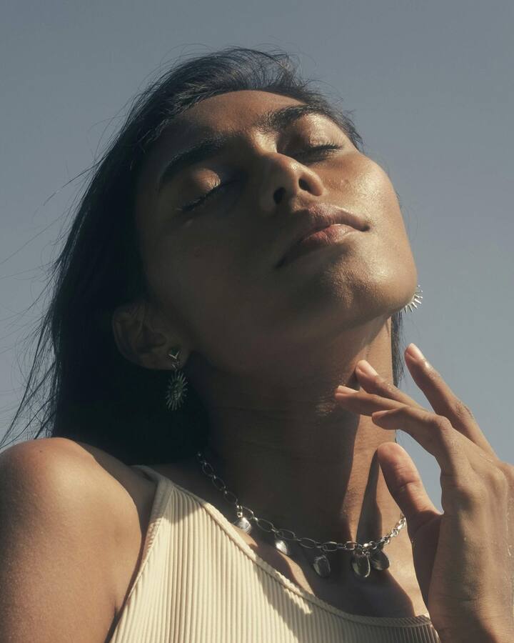 A woman with eyes closed tilts her face up to the sun, one hand resting at her collarbone, against a clear sky.