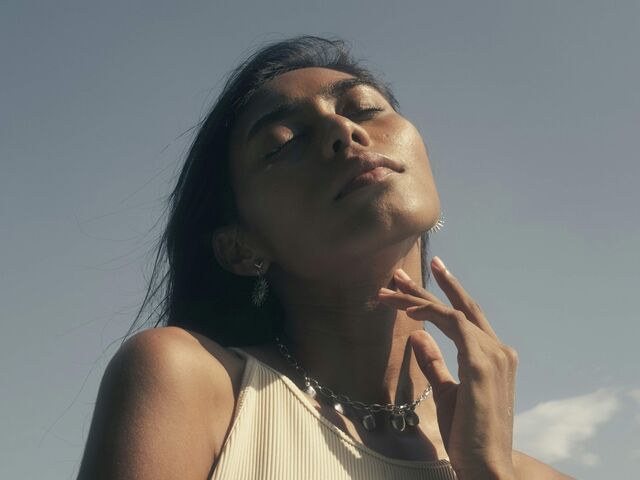 A woman with eyes closed tilts her face up to the sun, one hand resting at her collarbone, against a clear sky.
