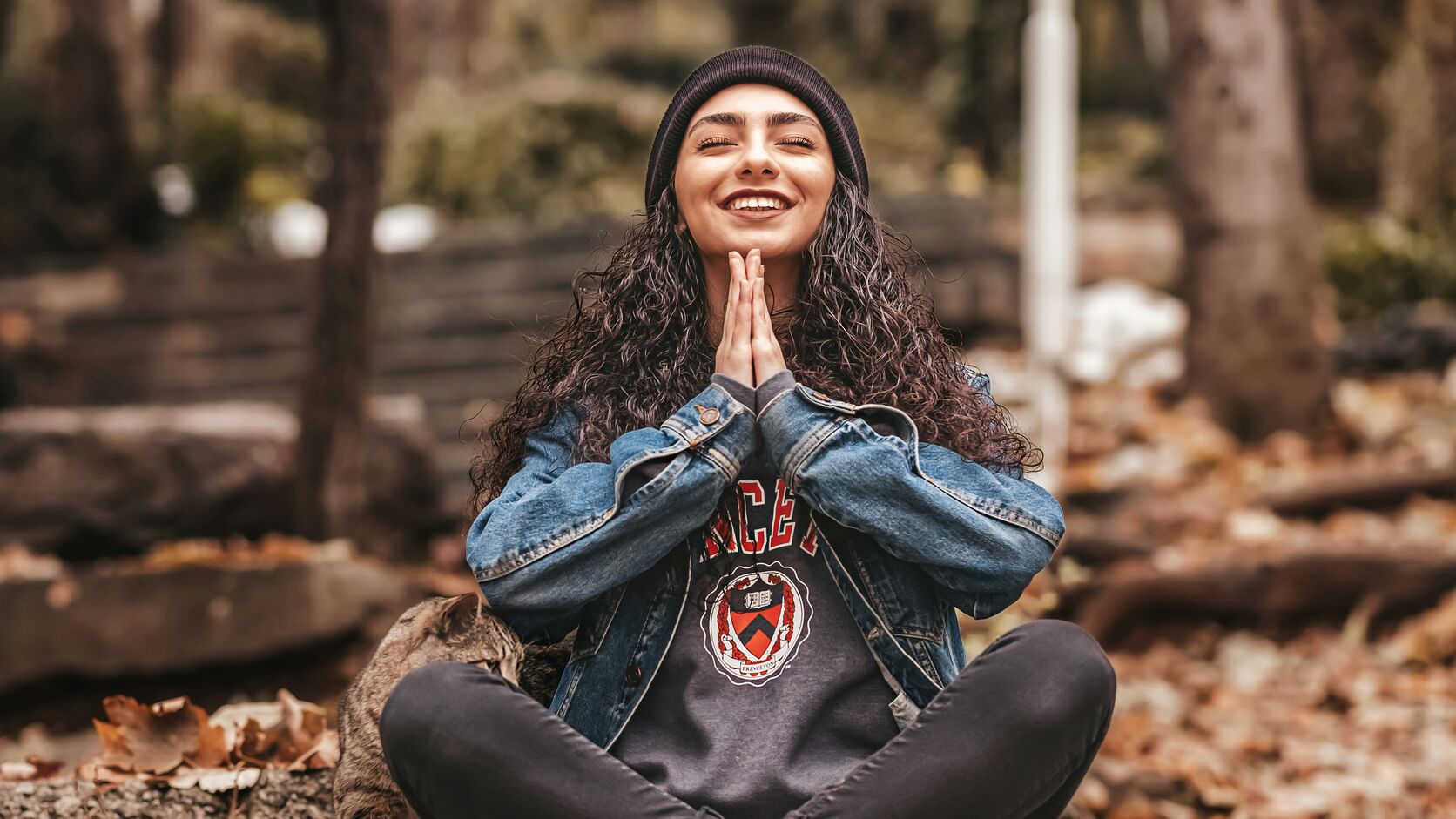 A smiling woman in a black beanie and denim jacket sits cross-legged on fallen autumn leaves, eyes closed and palms pressed together, a tabby cat at her side.