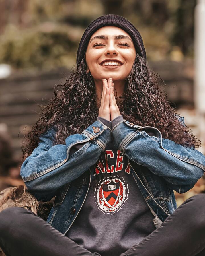 A smiling woman in a black beanie and denim jacket sits cross-legged on fallen autumn leaves, eyes closed and palms pressed together, a tabby cat at her side.