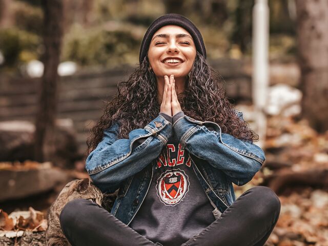 A smiling woman in a black beanie and denim jacket sits cross-legged on fallen autumn leaves, eyes closed and palms pressed together, a tabby cat at her side.