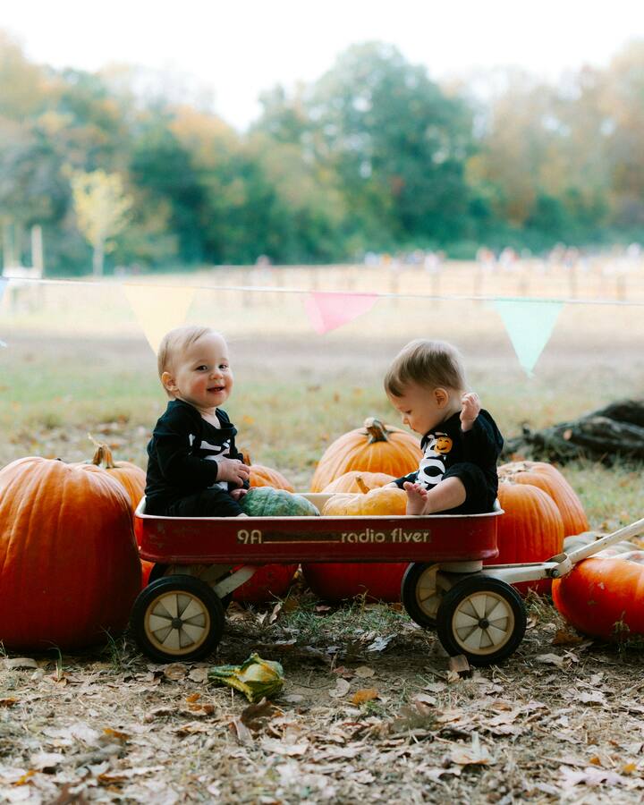 Two babies in black Halloween onesies sit in a red Radio Flyer wagon among orange pumpkins at a pumpkin patch, one grinning at the camera, with colorful bunting and autumn trees behind them.