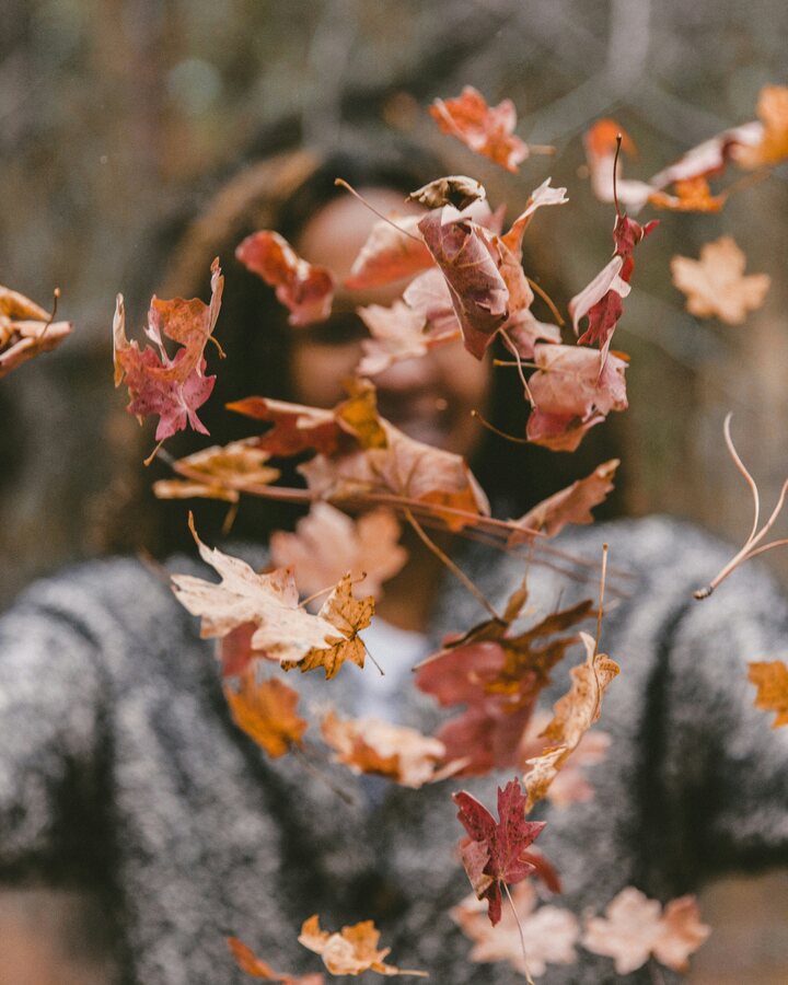 A woman in a grey knit sweater stands in an autumn wood with her arms flung wide as a flurry of red and orange maple leaves fills the air in front of her.