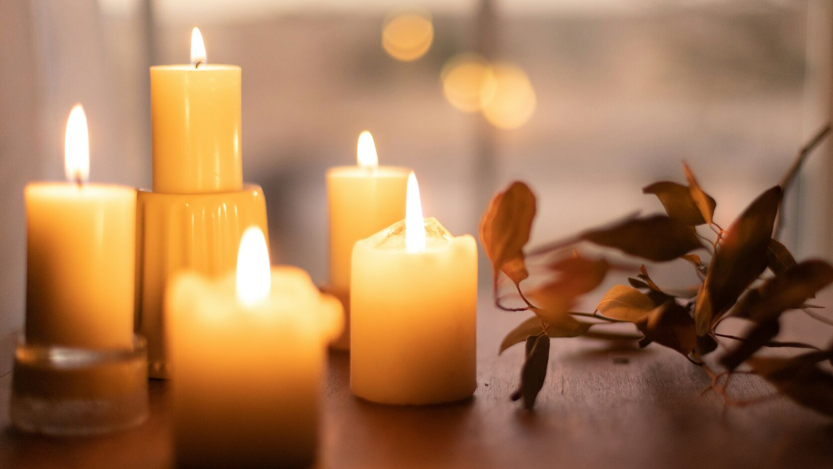 Five lit pillar candles glow on a wooden table by a window beside a sprig of dried autumn leaves, with soft golden bokeh in the window behind them.