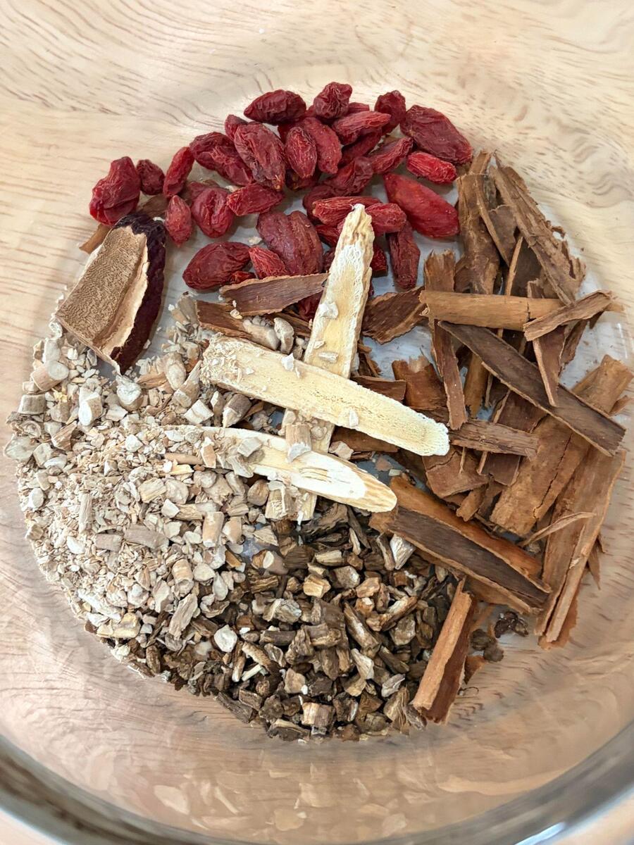 Decoction herbs in a glass bowl: dried goji berries, a slice of reishi mushroom, broken cinnamon bark, pale strips of astragalus root, and small pieces of ashwagandha and roasted burdock root.