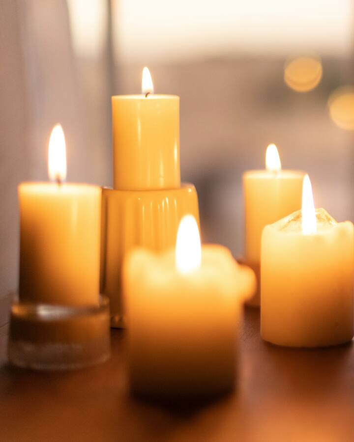 Five lit pillar candles glow on a wooden table by a window beside a sprig of dried autumn leaves, with soft golden bokeh in the window behind them.