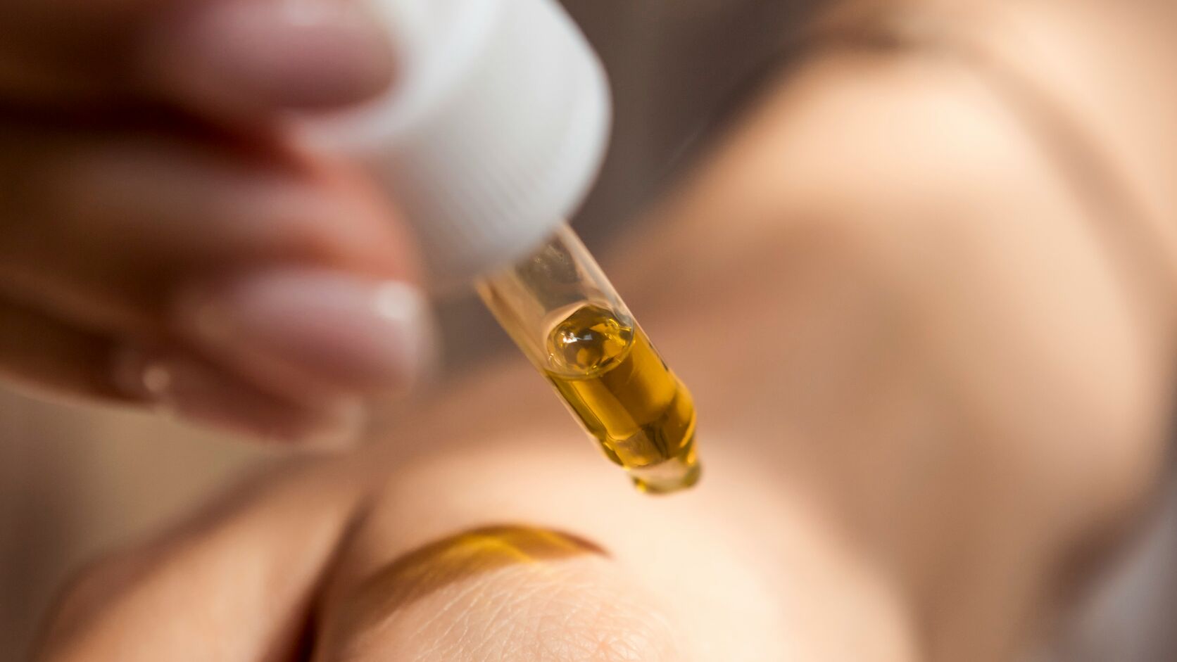 A hand with a pale mauve manicure squeezes a glass dropper of golden facial oil above the back of another hand, where a streak of oil already glistens.
