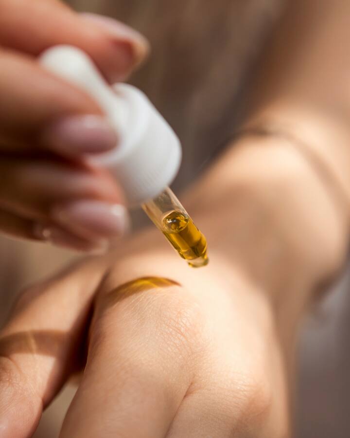 A hand with a pale mauve manicure squeezes a glass dropper of golden facial oil above the back of another hand, where a streak of oil already glistens.